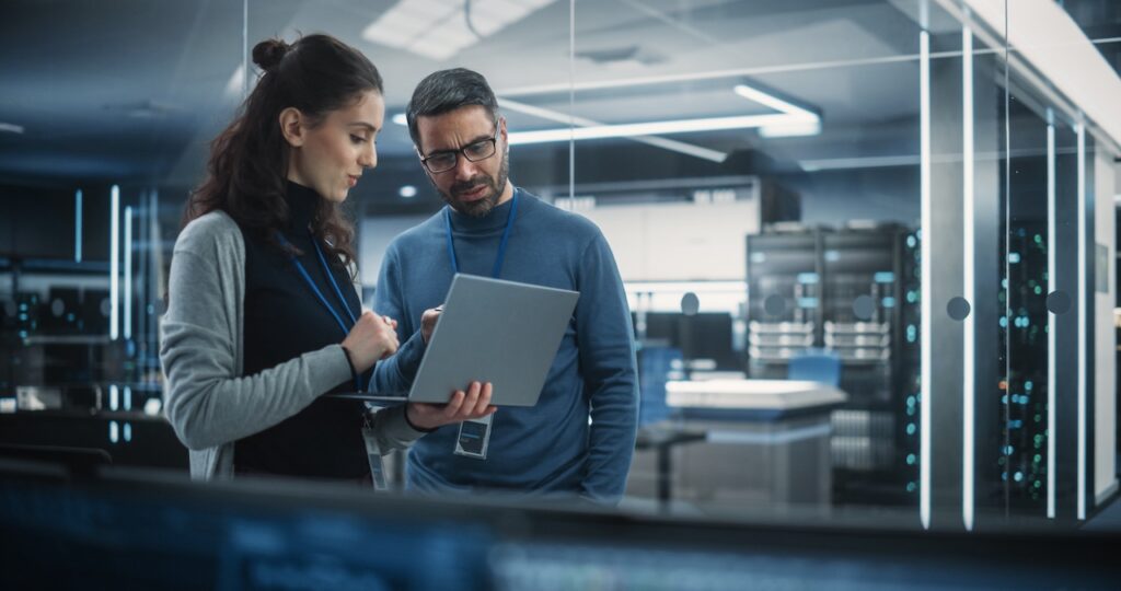 Zwei IT-Fachkraefte mit Ausweisen analysieren Daten auf einem Laptop in einem Serverraum
